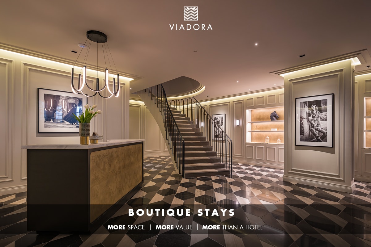 An elegantly designed lobby features a striking staircase with a modern light fixture above. A sleek reception desk, adorned with greenery, is complemented by wall-mounted art displays and softly illuminated shelves, highlighting the sophisticated atmosphere of the space.