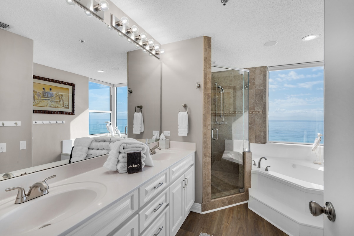 A bright bathroom features dual sinks with smooth, white countertops and ample storage. Large windows provide a view of the ocean, while a glass-enclosed shower and soaking tub add convenience. The space is accented with soft towels and modern lighting above the vanity.