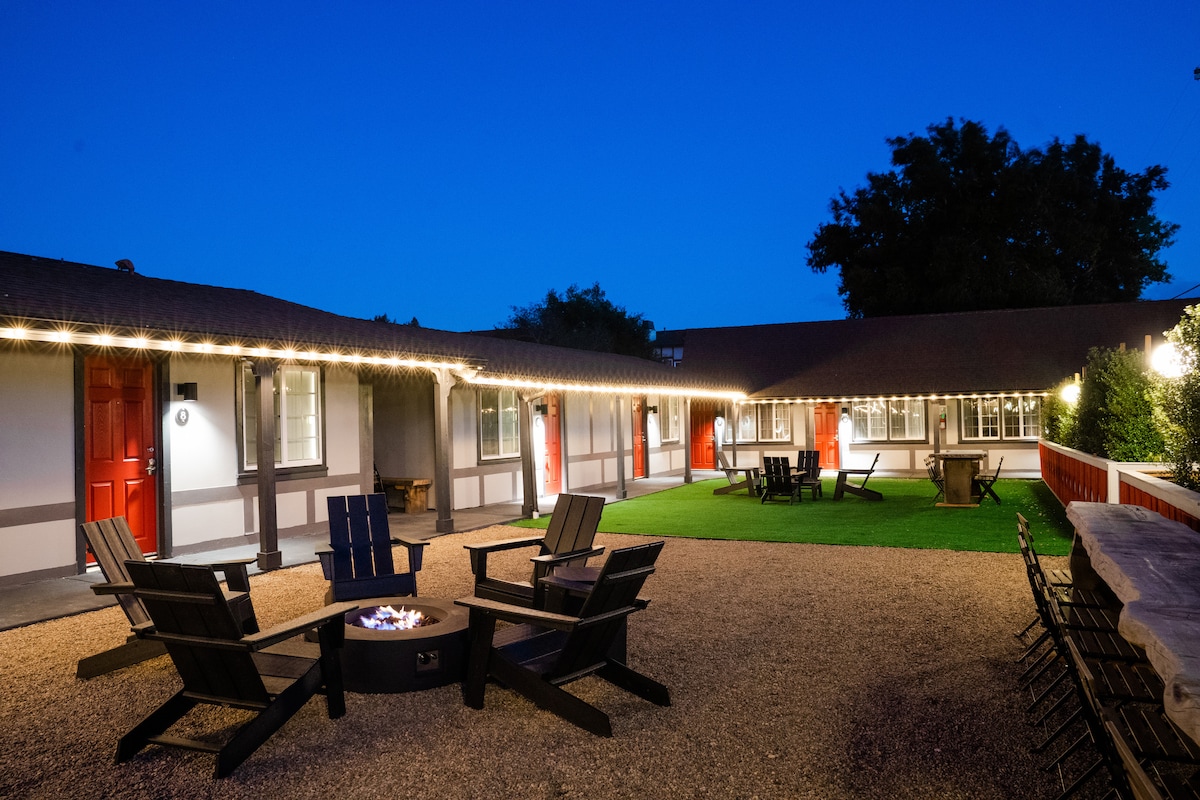 An outdoor gathering area features a fire pit surrounded by adirondack chairs, inviting relaxation. String lights illuminate the space, which is bordered by manicured grass and rustic stone paths. The motel's exterior is visible, showcasing red doors and large windows.
