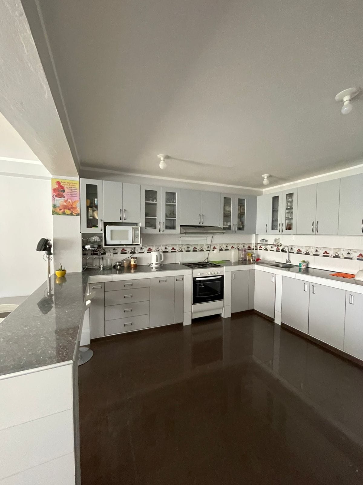 A spacious kitchen is equipped with white cabinetry and modern appliances, including an oven and microwave. A marble countertop surrounds the area, with ample storage provided by upper cabinets. Bright lighting illuminates the space, highlighting the cleanliness and organization of the kitchen.