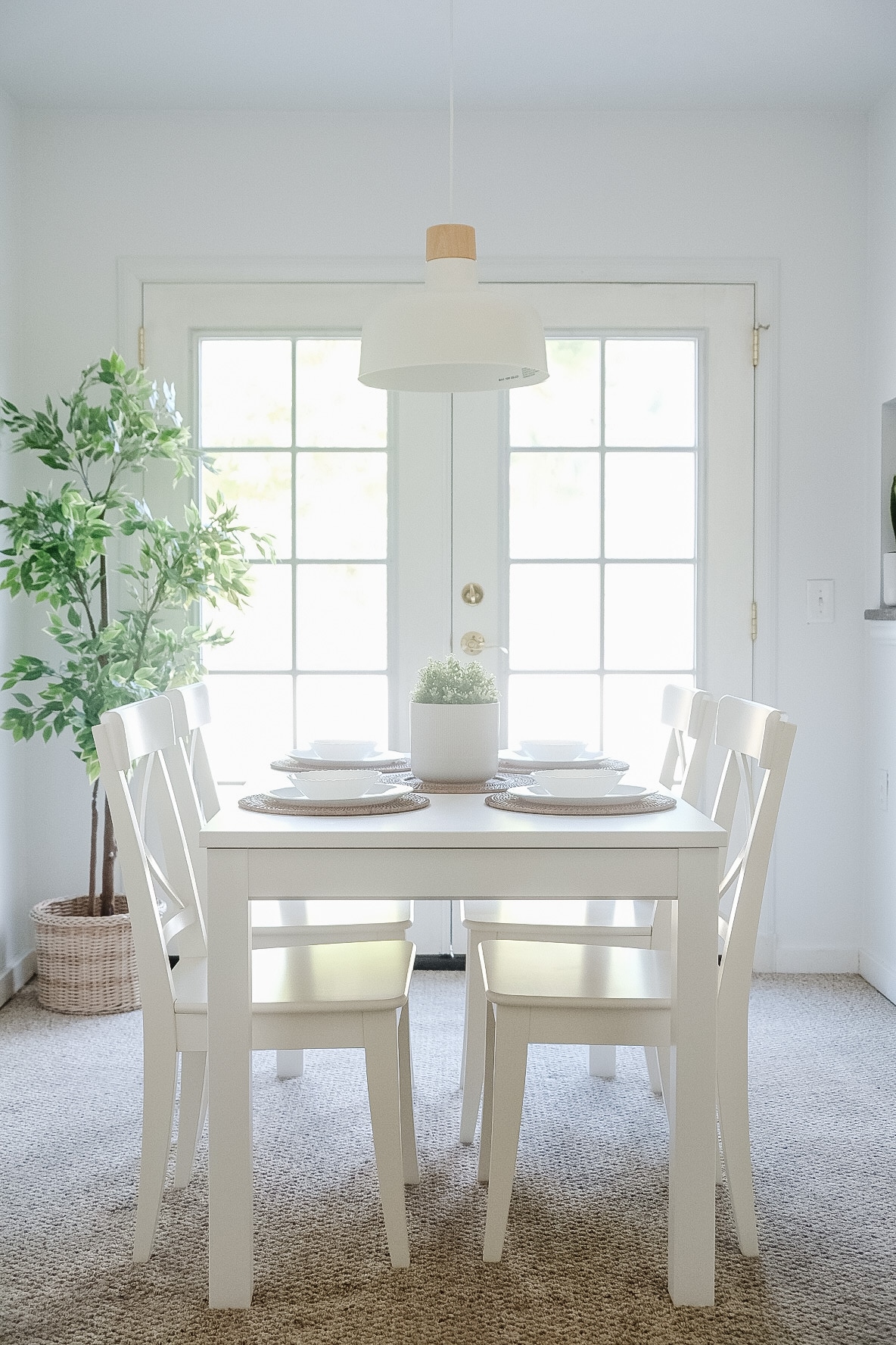 A bright dining area features a white table surrounded by four matching chairs. A simple centerpiece sits on the table, complemented by neutral table settings. A tall plant adds a touch of greenery, and natural light streams through the glass-paned doors.