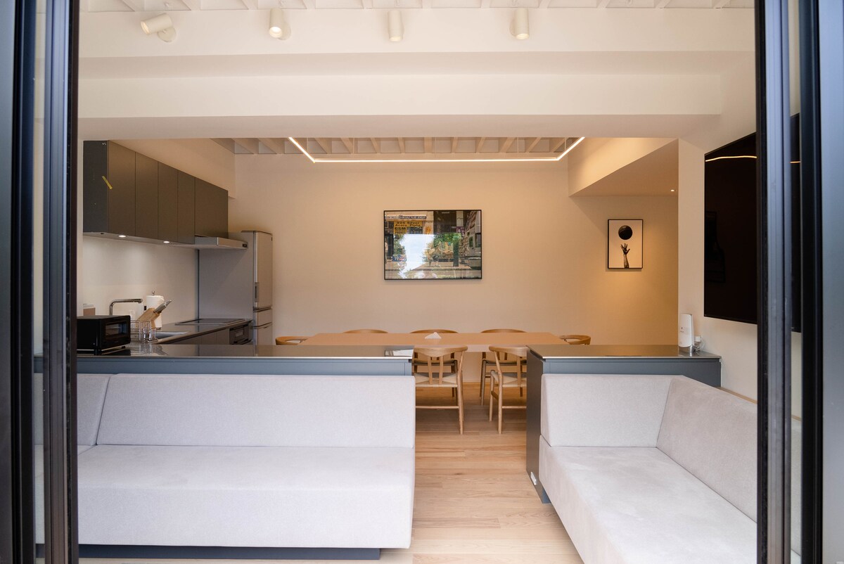 An open and airy common area features minimalist design elements with a spacious dining table surrounded by wooden chairs. A modern kitchen with sleek cabinetry and appliances is visible, enhancing functionality. Natural light floods the space through large glass doors, creating a warm ambiance.