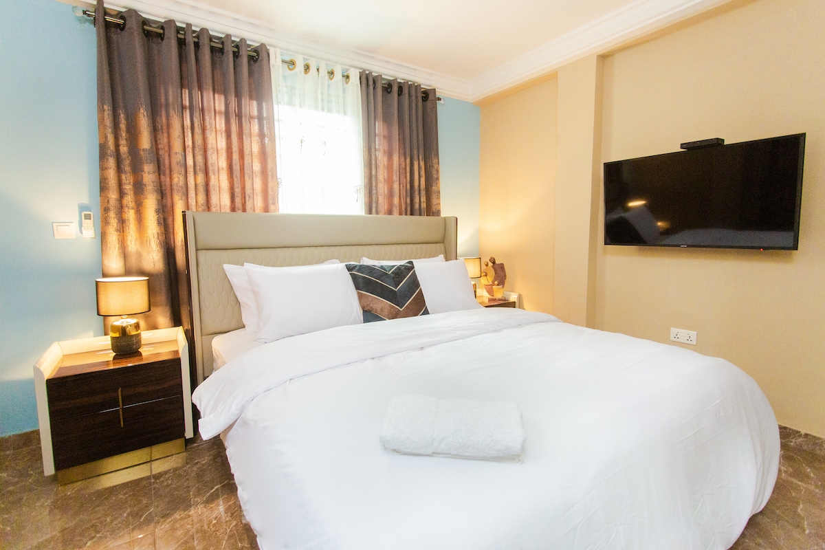 A well-appointed sleeping area features a spacious bed dressed in crisp white linens. Adjacent nightstands with lamps provide soft illumination, while large curtains frame the window, allowing natural light to filter gently into the room. A wall-mounted television is positioned for easy viewing.