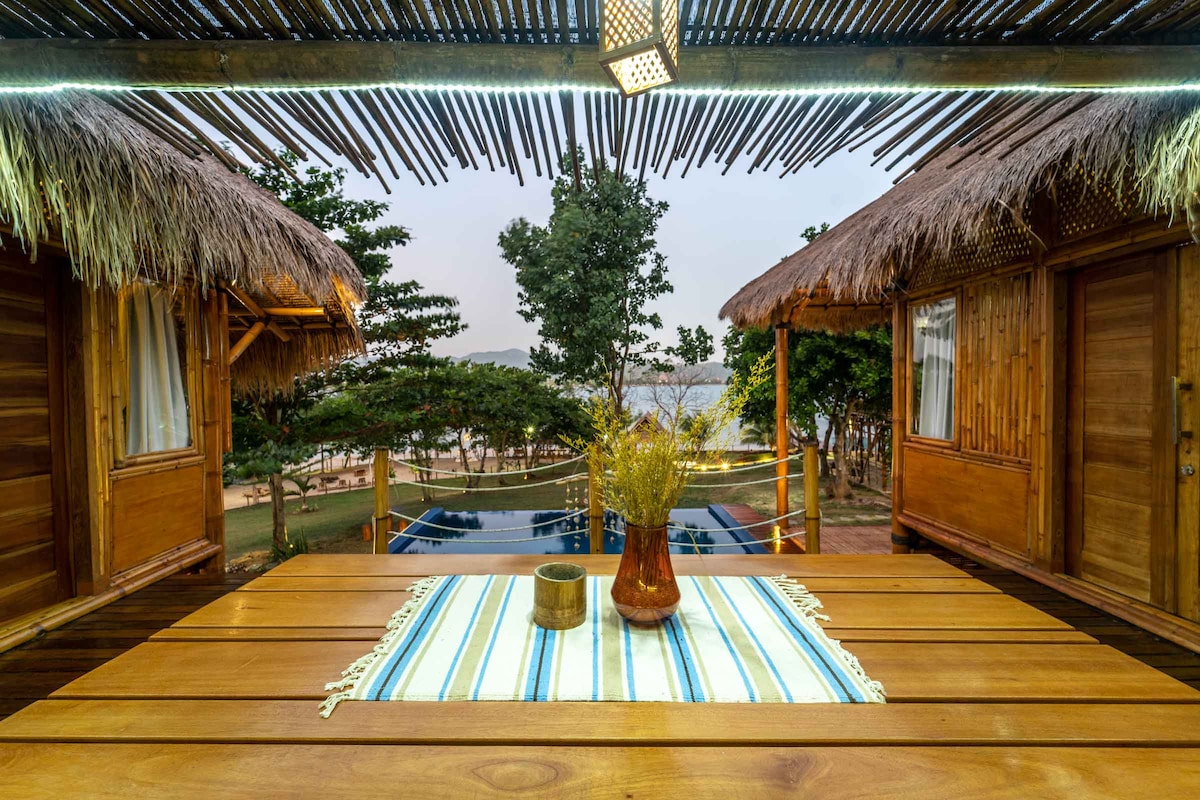 A wooden dining table is set under a thatched roof, featuring a vase with dried flowers and a striped table runner. Two cozy bungalows are visible in the background, with lush greenery surrounding a swimming pool, creating a serene atmosphere.
