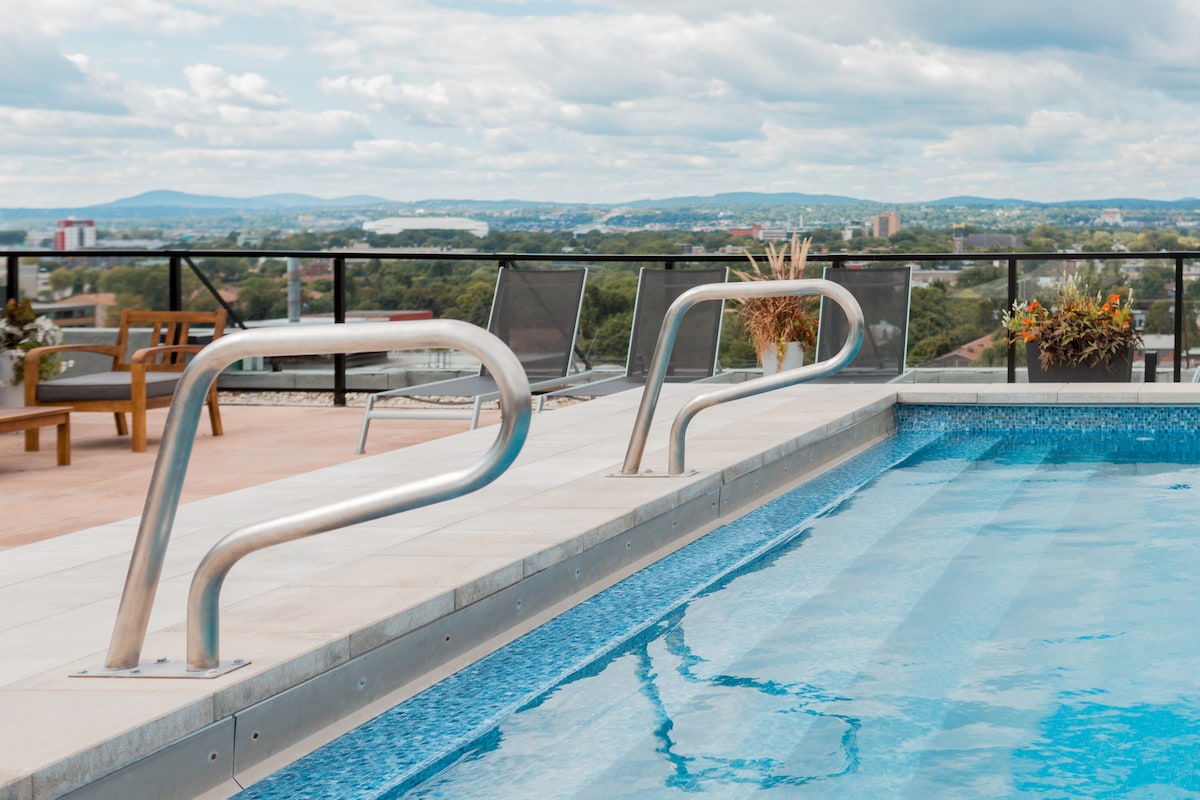 A rooftop pool features a clear blue water surface, with sleek stainless steel railings on either side. Comfortable lounge chairs are set along the edge, surrounded by potted plants and a scenic view of the city skyline in the distance.