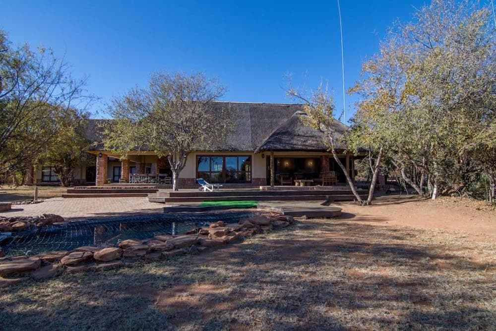 A spacious chalet is set against a clear blue sky, surrounded by trees and landscaped grounds. The exterior features a thatched roof, large windows, and a wide deck area. A green lawn extends towards the foreground, adding to the serene ambiance.