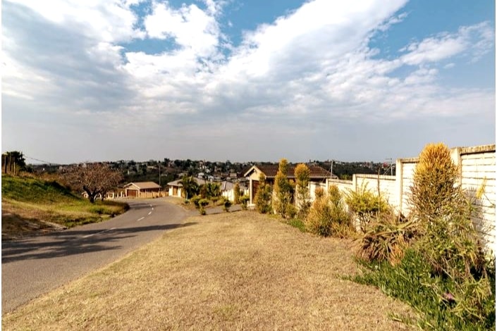 A well-maintained street view displays a gently sloping landscape with grass and scattered plants along the roadside. Residential homes are visible in the distance, framed by a clear sky with soft clouds, reflecting a peaceful neighborhood ambiance.