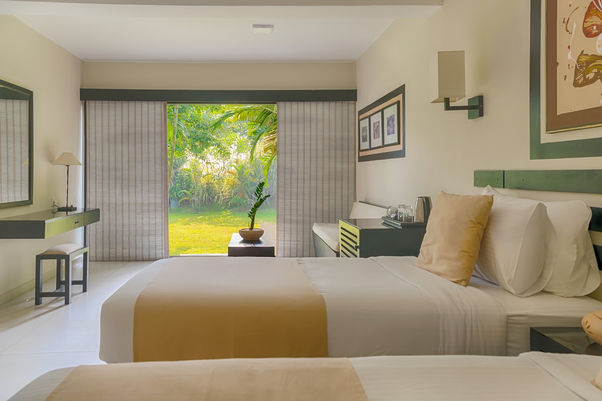 A bright and airy room features two spacious beds dressed in white linens. Large glass doors open to a serene outdoor view of a lush garden. A small writing desk and lamp are positioned against one wall, complemented by framed artwork and minimal furnishings.