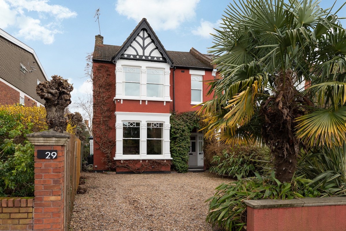 The Edwardian family home features a striking red facade with characteristic architectural details, framed by lush greenery. A large, gravel driveway leads to the entrance, flanked by mature tropical plants and trees, offering a welcoming first impression.