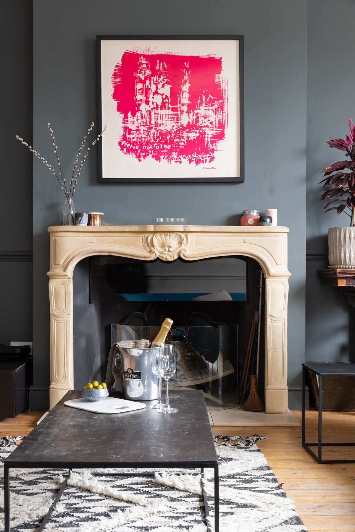 A classic stone fireplace is centered against a deep gray wall, adorned with an abstract artwork in vibrant pink. A stylish, low coffee table sits in front of the fireplace, accompanied by elegant glassware and a bottle of champagne in an ice bucket. Decorative plants enhance the ambiance.