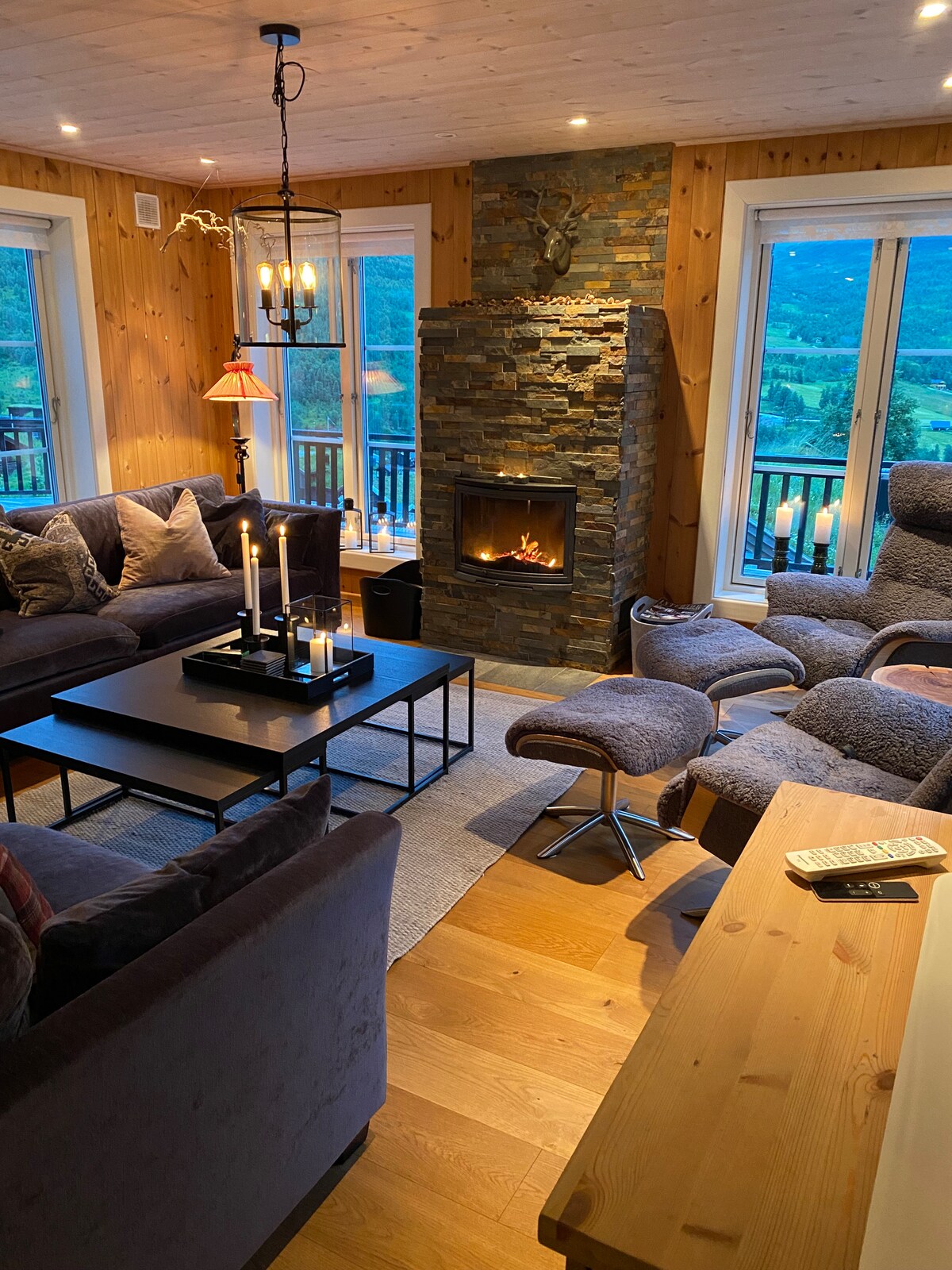 A cozy living area is featured, with large windows providing views of the surrounding mountains. A stone fireplace is centered among comfortable seating, including plush armchairs and a sectional sofa. A coffee table is arranged with candleholders, creating a relaxed and inviting ambiance.