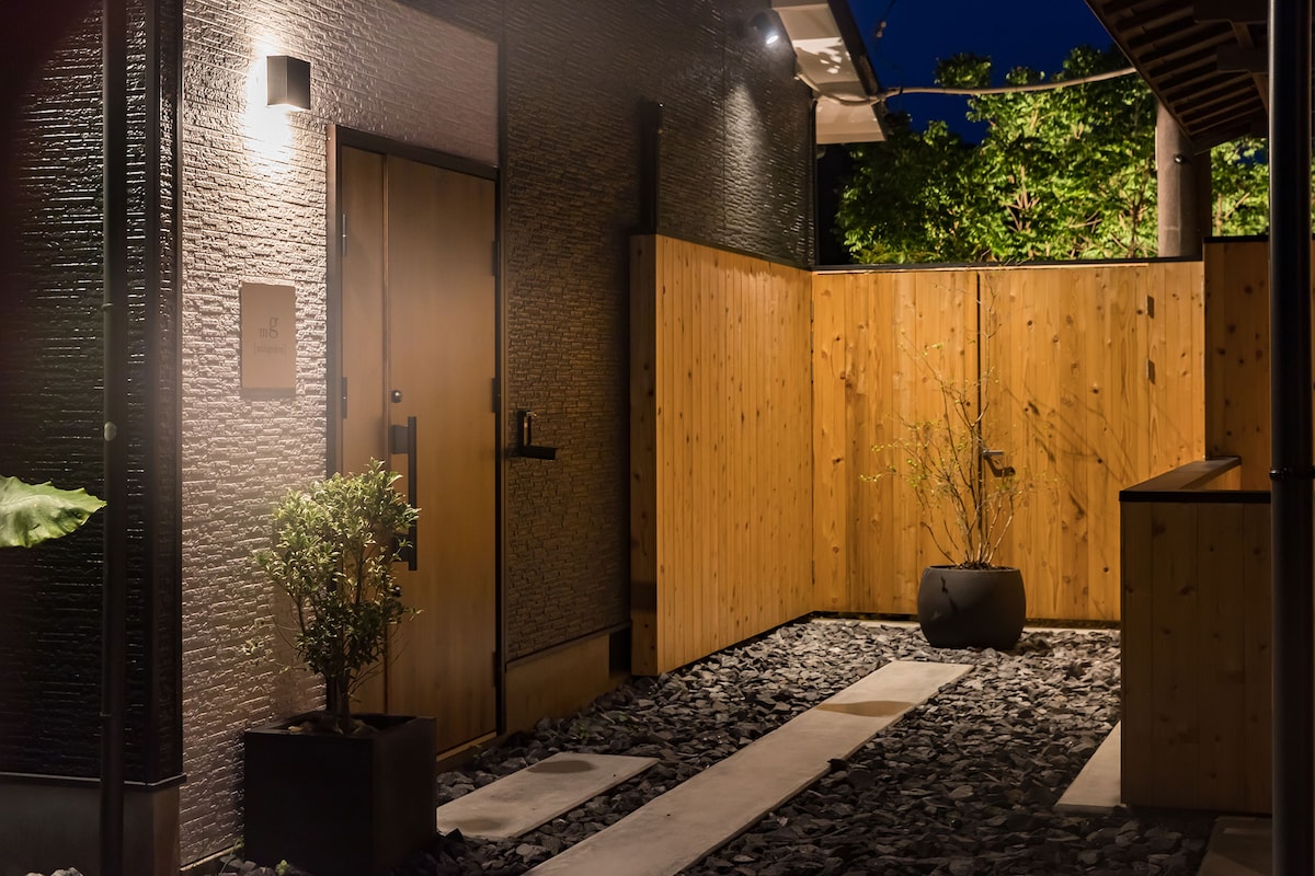 A serene entrance is framed by a combination of textured dark walls and warm wooden fencing. A stone path leads to the door, complemented by subtle outdoor lighting. Lush greenery is visible in the background, enhancing the tranquil atmosphere.