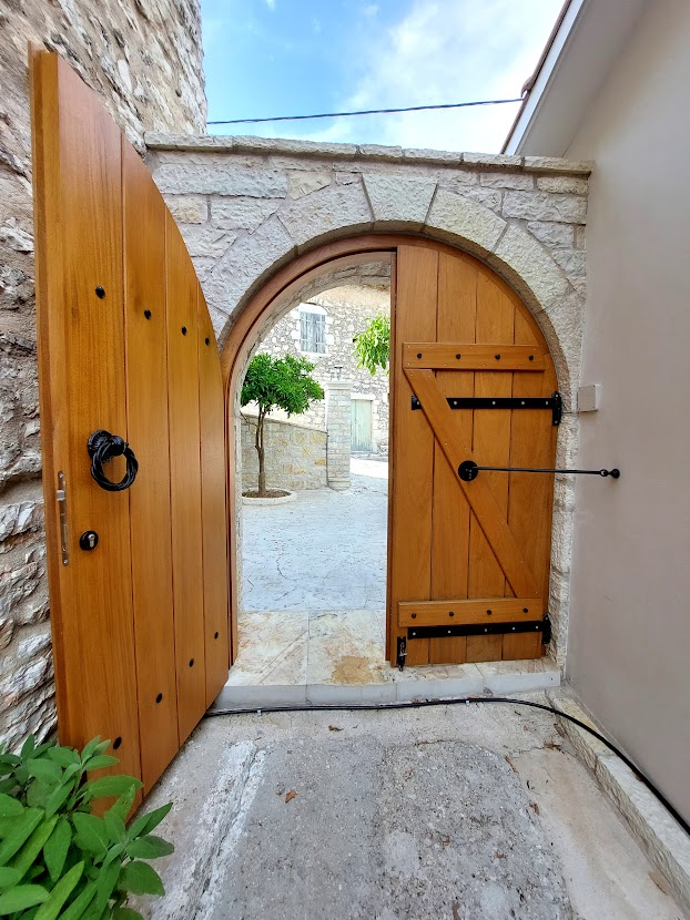 An entrance features a large, wooden arched door with a decorative metal handle. The door is partially open, revealing a stone courtyard adorned with greenery. Sunlight filters through the opening, illuminating the surrounding stone walls.