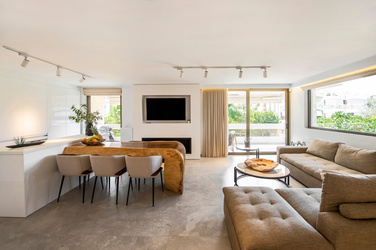 A spacious living area is presented with large windows offering natural light. A modern sectional sofa faces a flat-screen television mounted on the wall. A wooden dining table with soft seating is positioned nearby, with greenery visible outside through the glass doors.