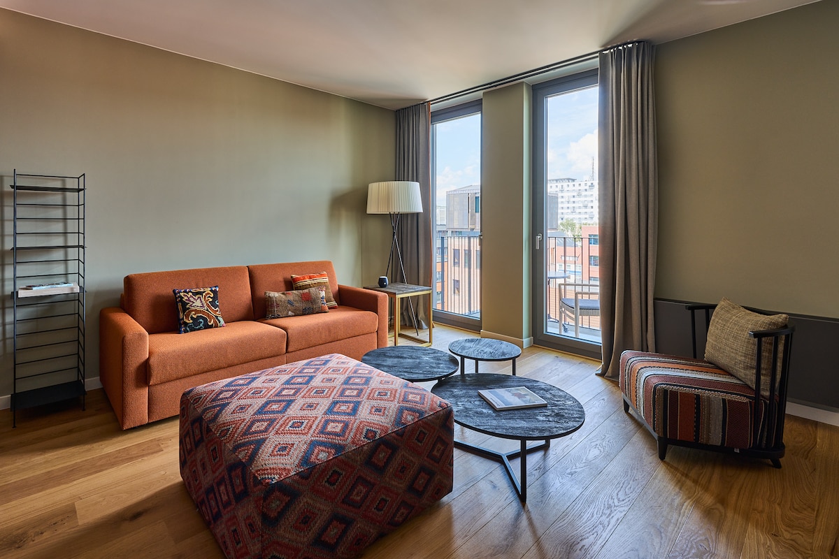 A spacious living area is highlighted by a bright orange sofa and a patterned ottoman. Two round coffee tables with dark surfaces are arranged in front, while tall windows invite natural light. A floor lamp stands beside the sofa, complementing the neutral wall tones.