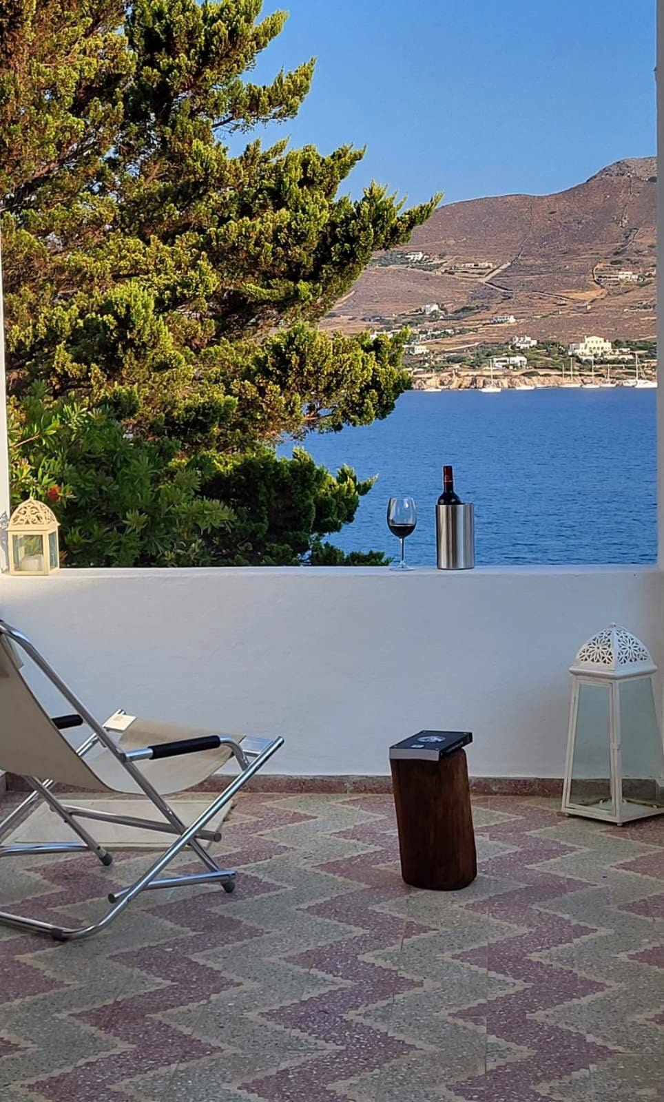 A cozy outdoor seating area is presented with a folding chair positioned on a patterned tiled floor. A small wooden table holds a wine bottle and glass. Scenic views of the sea and hills are visible in the background, framed by lush greenery.