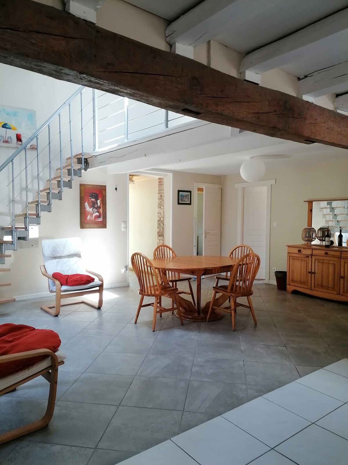 An open living space features a wooden dining table surrounded by four chairs. A staircase leads to a second level, while a comfortable chair with a cushion is positioned nearby. Natural light enters through windows, highlighting the neutral color palette and textured walls.