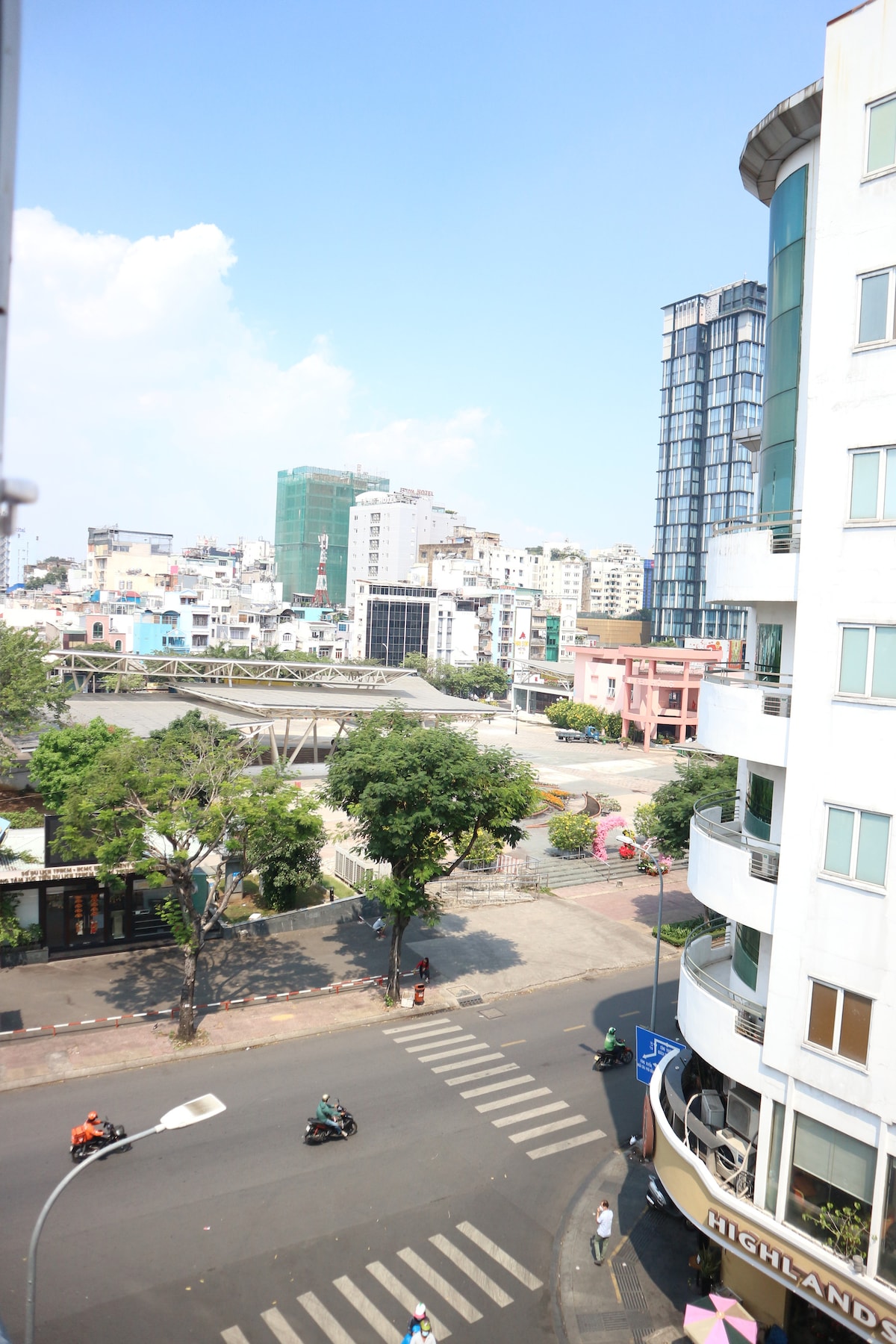 A cityscape view showcases a blend of architectural styles, with tall modern buildings and a mix of colorful structures in the background. A street crosses in front, featuring occasional vehicles and cyclists. Lush trees provide greenery amidst the urban environment.