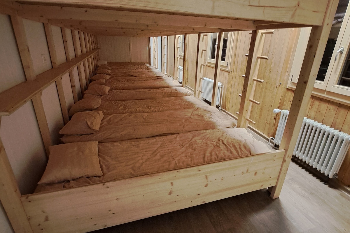 A spacious dormitory area is featured, showcasing six individual sleeping spaces arranged on elevated wooden platforms. Each platform is equipped with mattresses and pillows, providing a simple yet functional design. The room is framed by wooden walls and multiple windows allowing natural light.