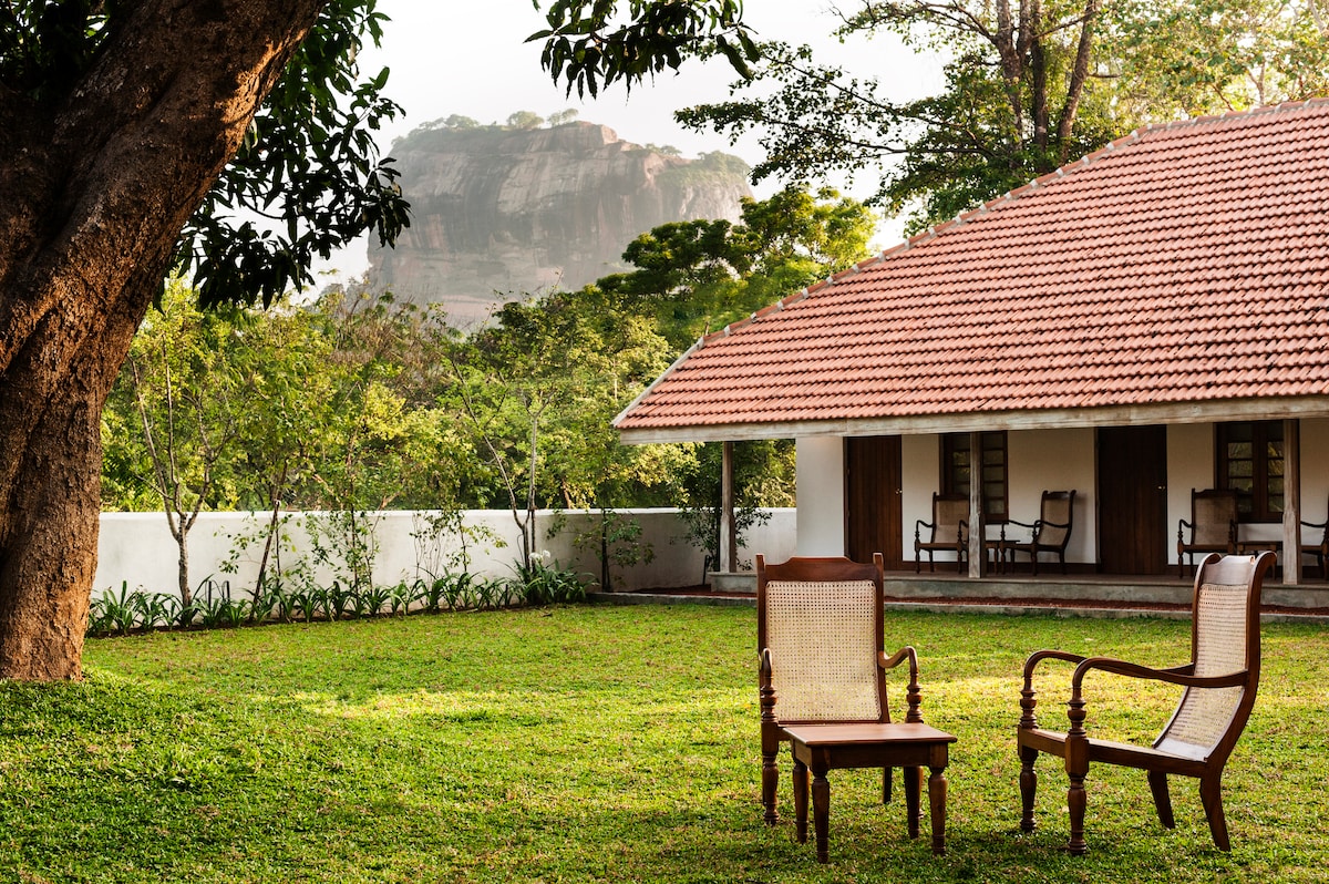 A serene outdoor space features two wooden chairs positioned on a grassy lawn. A traditional structure with a red-tiled roof and wooden doors can be seen in the background, framed by lush greenery. The prominent Sigiriya rock formation is visible beyond the property.