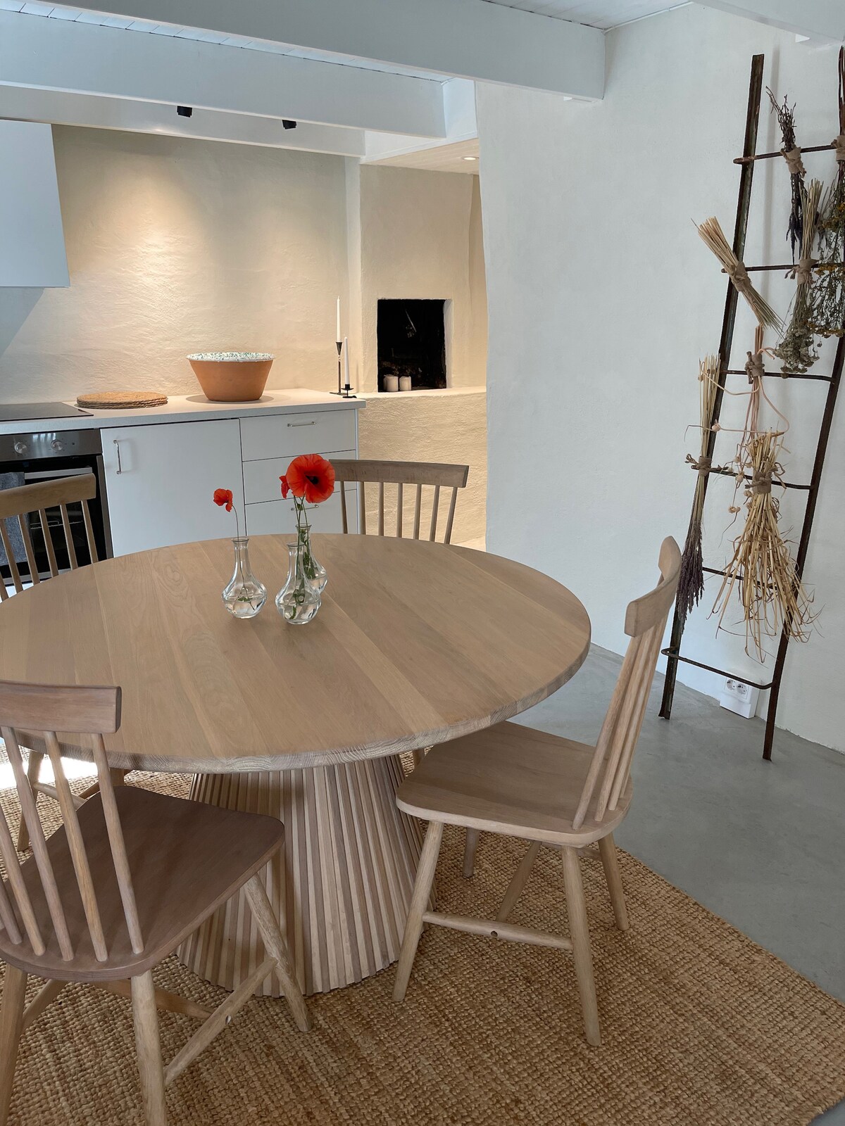 A round wooden dining table is placed at the center, accompanied by two matching chairs. A simple centerpiece with two vases holding red flowers adds a touch of color. The background features a minimalist kitchen with a light-colored wall, enhancing the overall airy feel.