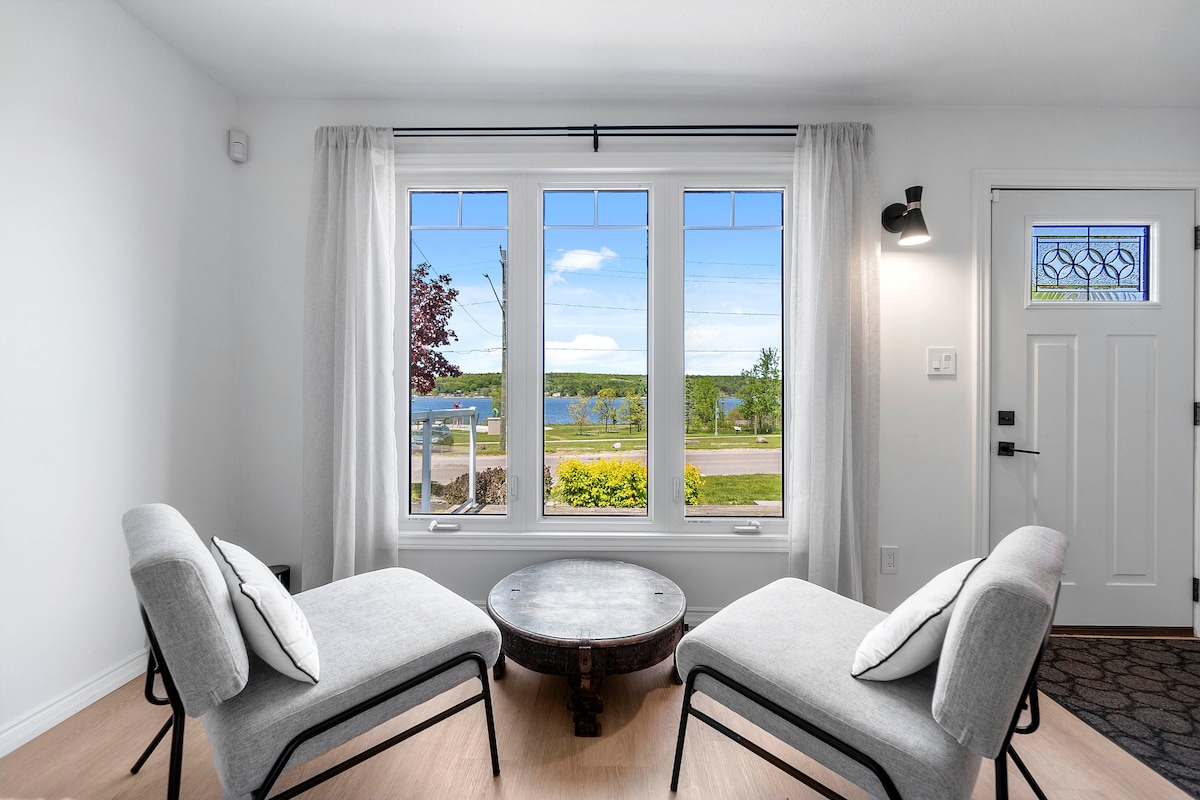Two comfortable chairs are positioned across from a round coffee table, offering a serene view through large windows. The scene includes a waterfront vista with greenery and pathways visible, framing an inviting atmosphere filled with natural light.