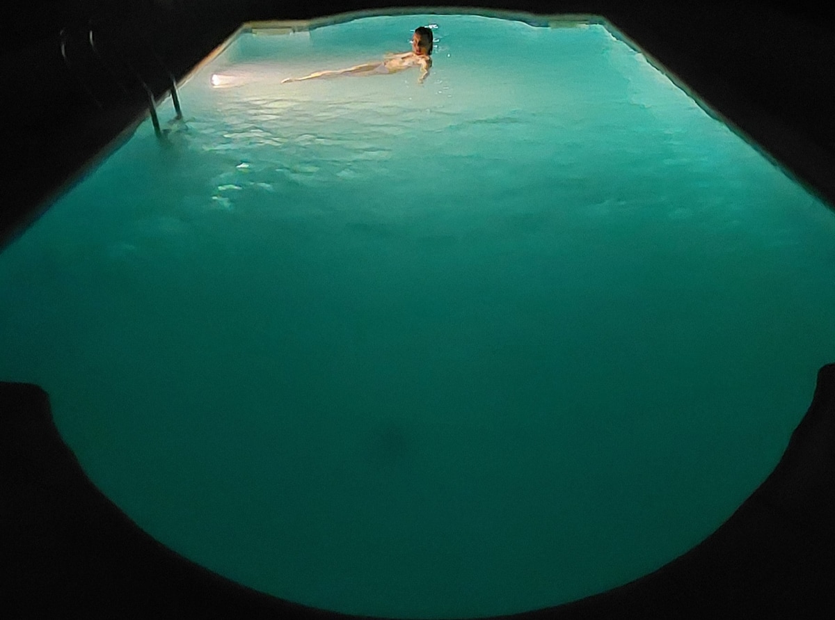 A serene swimming pool is illuminated in soft turquoise light, creating a calm ambiance. A figure is seen gently floating on the surface, emphasizing the inviting nature of the pool. The surrounding area remains dark, enhancing the tranquility of the setting.