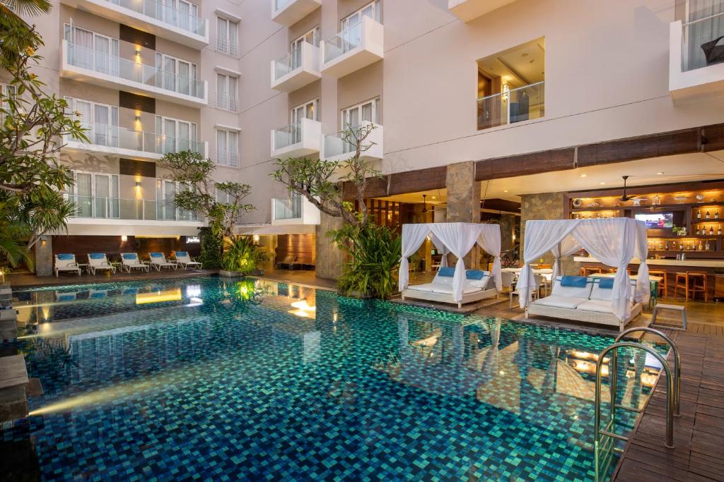 A tranquil swimming pool is surrounded by lush greenery and comfortable lounge beds with canopies. The pool's vibrant blue tiles reflect the light, while a bar area is visible in the background. The modern building features large balconies that overlook the serene space.