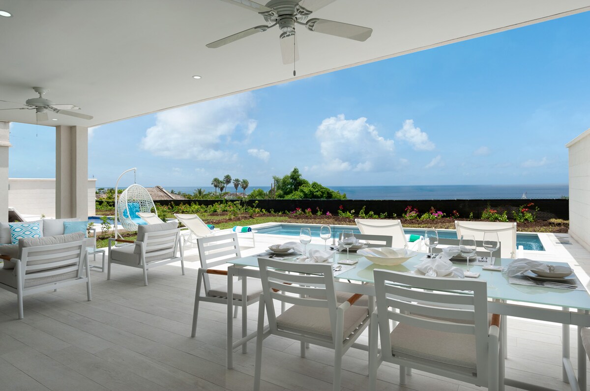 An expansive outdoor terrace features a dining table set with tableware, surrounded by comfortable seating. A private pool is visible, framed by colorful tropical plants. The serene ocean view and clear blue sky enhance the inviting space, perfect for relaxation and enjoyment.