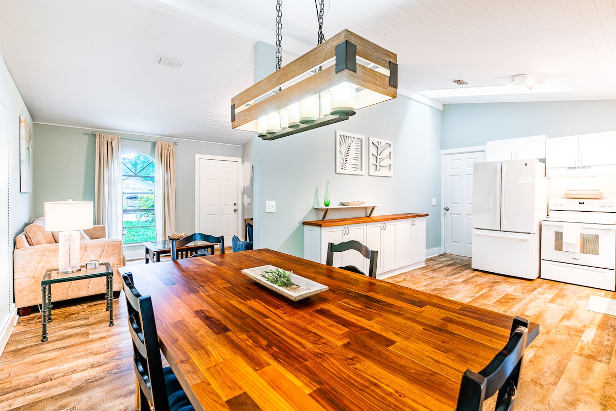 A spacious dining area features a large wooden table surrounded by black chairs, complemented by a modern light fixture above. A cozy seating area with a beige couch is visible. Bright natural light enters through window treatments, enhancing the inviting atmosphere of the open layout.