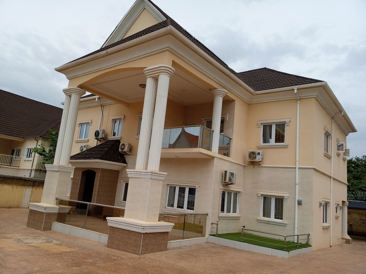 A modern two-story building is showcased, featuring a light-colored exterior with elegant architectural details, including columns and a balcony. Multiple air conditioning units are visible, and a well-maintained outdoor area is presented with a grassy section. Large windows ensure ample natural light.
