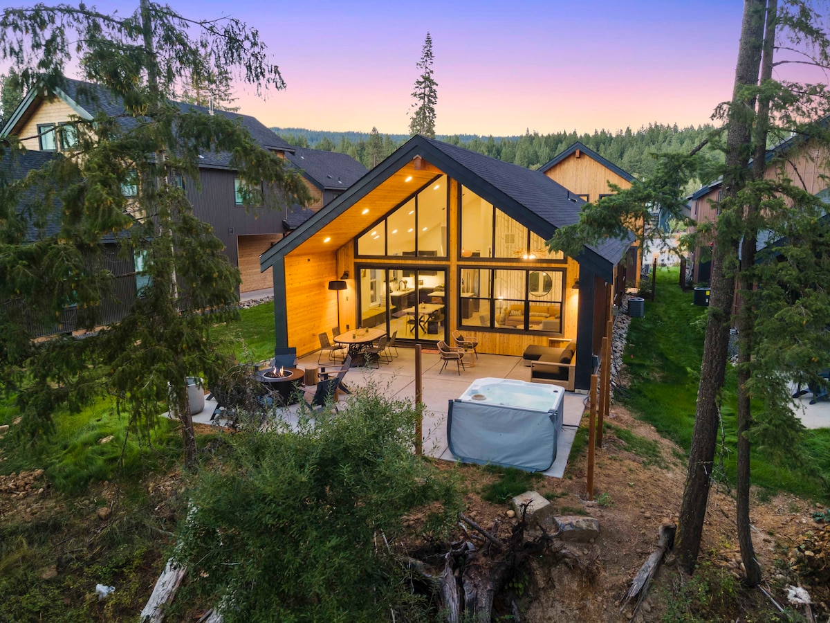 A modern cabin is viewed from above, featuring large glass windows that allow natural light to illuminate the interior. A spacious back patio includes a hot tub and seating area, set within a forested landscape with seasonal greenery.