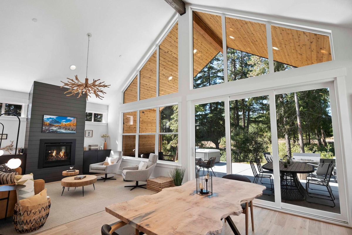 A spacious living area features large floor-to-ceiling windows allowing natural light to flood the room. The modern design includes a fireplace, comfortable seating, and a dining table, providing a seamless connection to the outdoor patio with views of the surrounding nature.