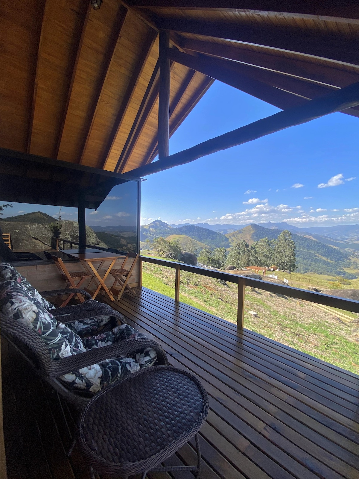 A covered balcony features two woven chairs and a folding table, providing a space to relax. The stunning view showcases rolling hills and distant mountains under a clear blue sky.