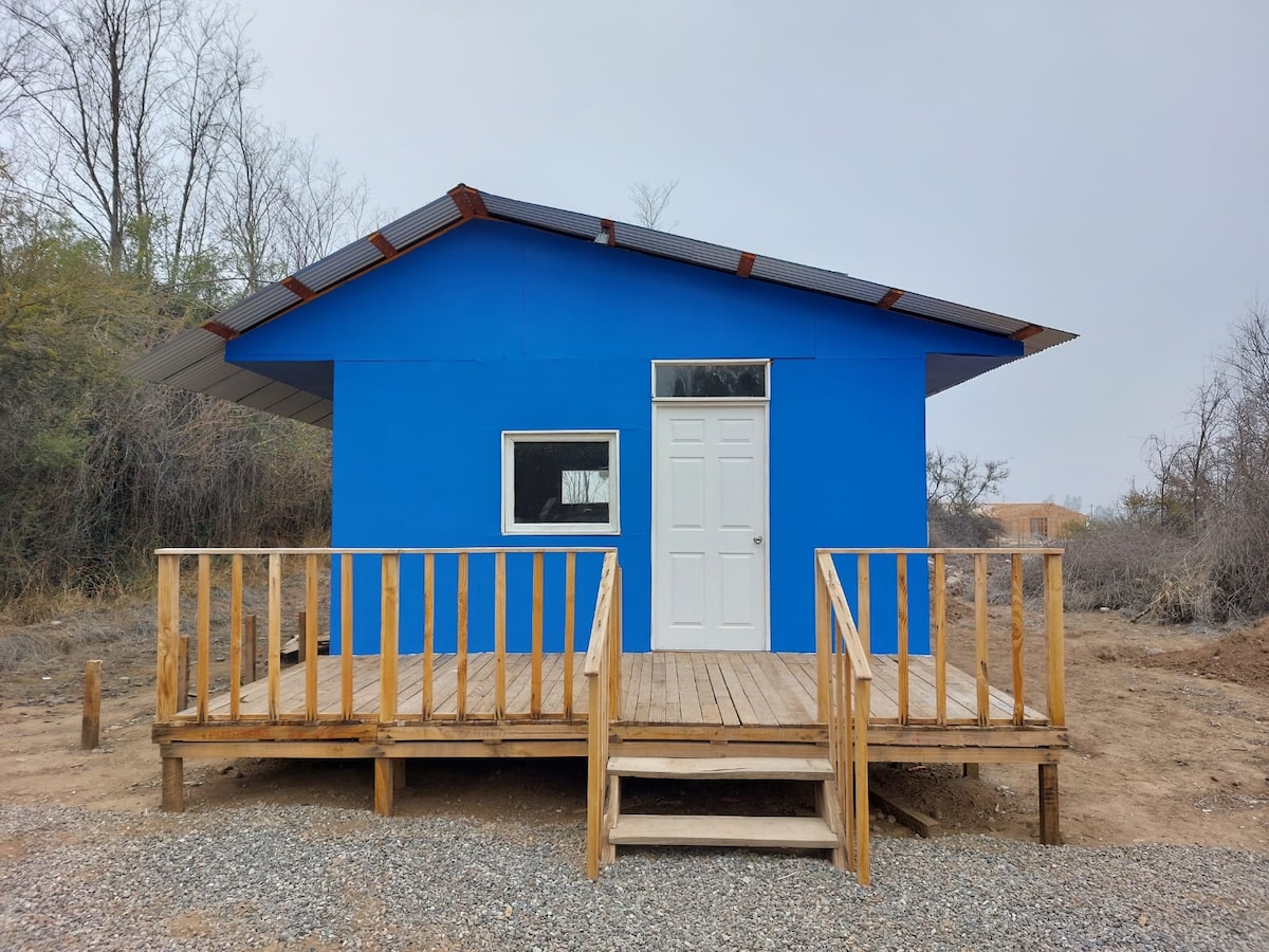 A bright blue structure is featured, set on a wooden deck with steps leading to the entrance. A large window allows natural light to enter, while the simplistic design is surrounded by a natural landscape with sparse vegetation.