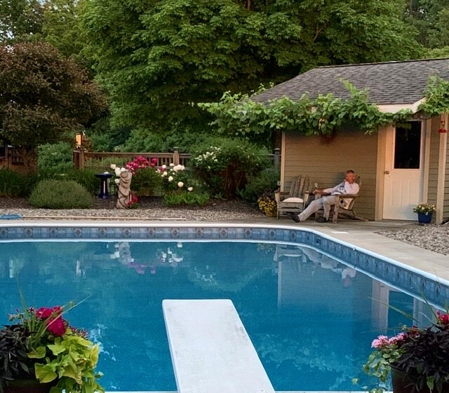 A serene pool area is framed by vibrant flower beds filled with various plants. A diving board extends over the clear water, while a cozy seating area with a person relaxing is positioned nearby under the shade of lush greenery. A garden shed can be seen in the background.