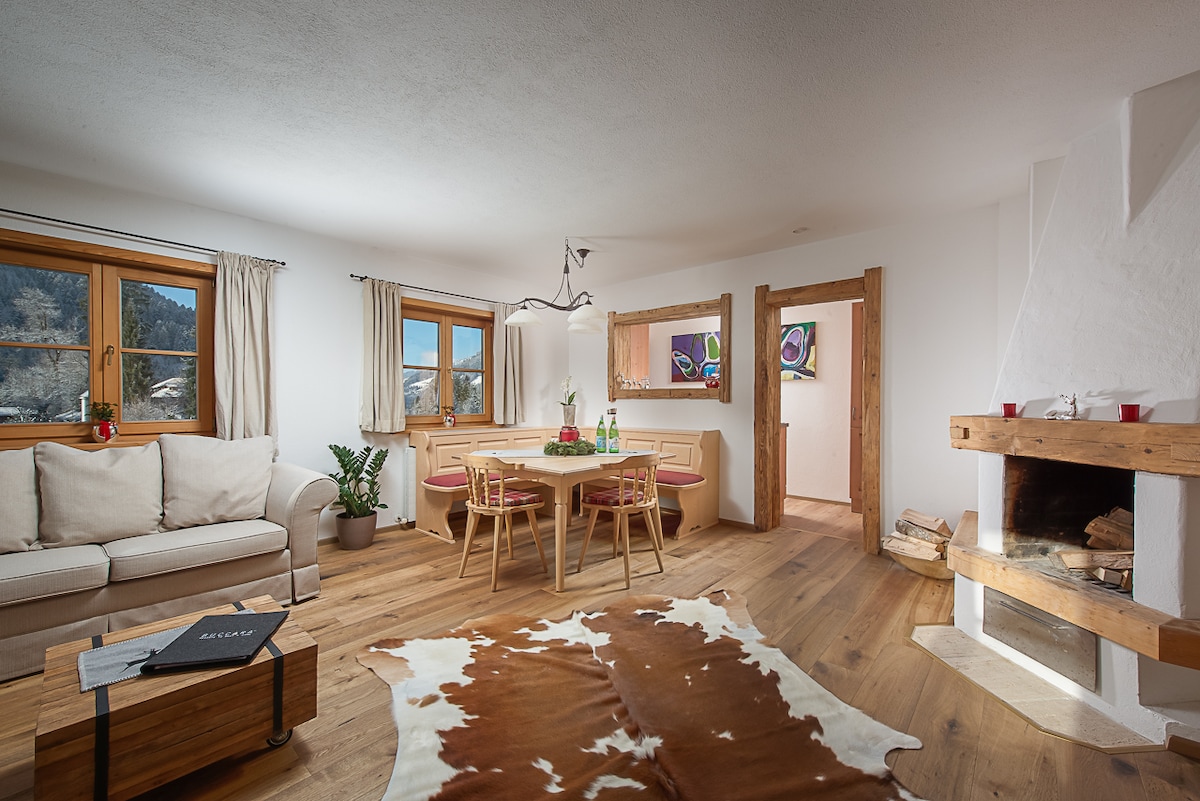 A cozy living area features a light-colored sofa and a wooden dining table surrounded by four chairs. A fireplace with wood storage is present, along with a decorative area rug. Large windows allow natural light to brighten the space, while mountain views are visible.