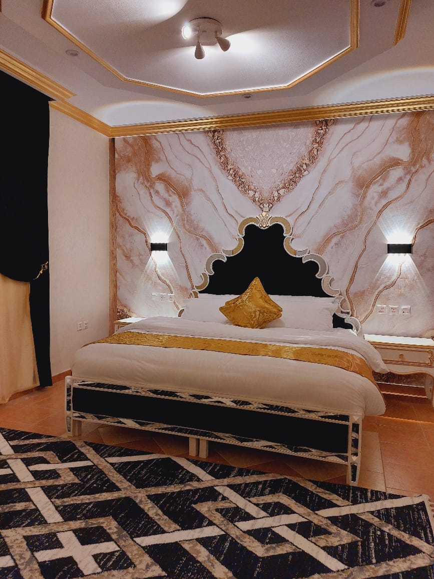 A spacious bedroom is highlighted by an elegant headboard featuring a distinctive marble design. A plush bed, adorned with decorative pillows, is complemented by stylish bedside lighting. A patterned rug adds texture to the room, while dark curtains frame the window for a cozy ambiance.