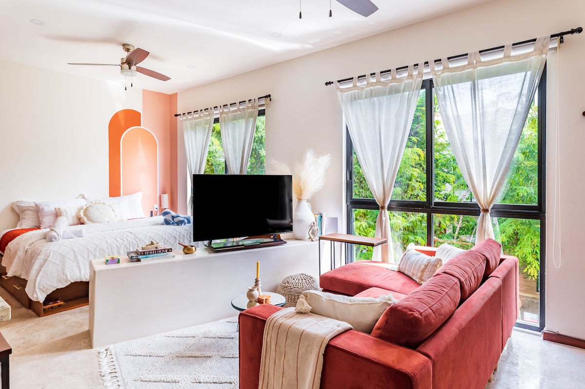 A spacious studio features large windows that invite natural light and offer views of lush greenery. A comfortable king-sized bed is situated against a softly colored wall, while a cozy seating area with a red sectional sofa and a TV occupies the opposite side.