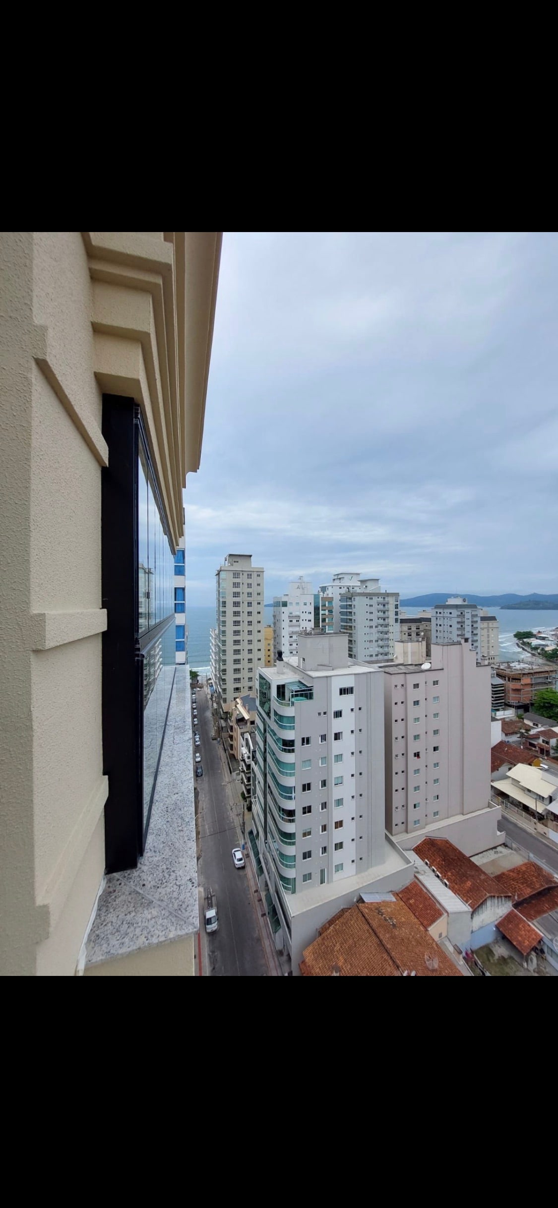An elevated view displays a blend of modern architectural structures, including high-rise buildings and rooftops. The street runs below, leading toward a coastal area visible in the distance, with a cloudy sky overhead providing a subdued light.