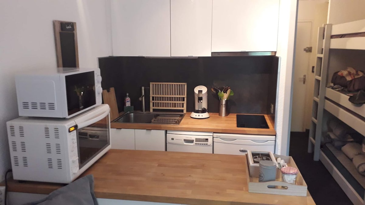 The kitchen area features sleek cabinets and a countertop made of light wood. Included are a microwave, oven, and a dishwasher. A Senseo coffee machine and cooking utensils are present. The open layout allows for easy movement between the kitchen and the living space.