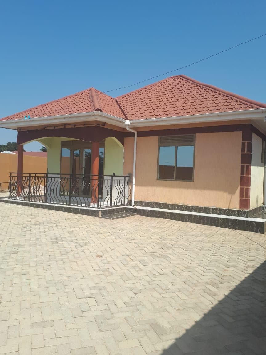 A single-story house features a red-tiled roof and a framed balcony. The exterior walls are a mix of light yellow and beige, complemented by dark stone trim. A clear blue sky serves as a backdrop, enhancing the home's inviting appearance.