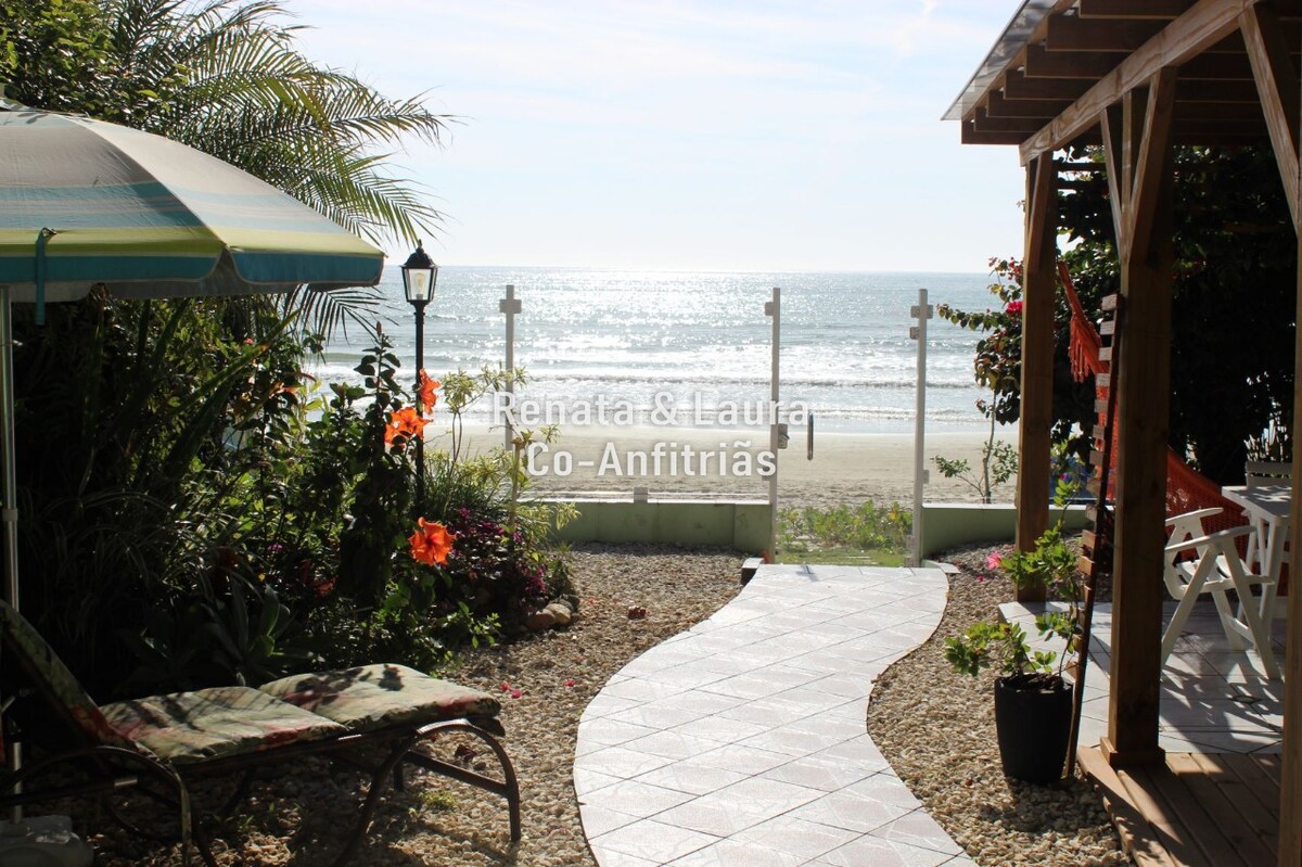 A tranquil outdoor area features a pathway leading towards the beach, surrounded by tropical plants and flowers. A shaded seating area with striped umbrellas is visible, along with a pergola that provides a cozy space for relaxation. Stunning ocean views are framed by a decorative fence.
