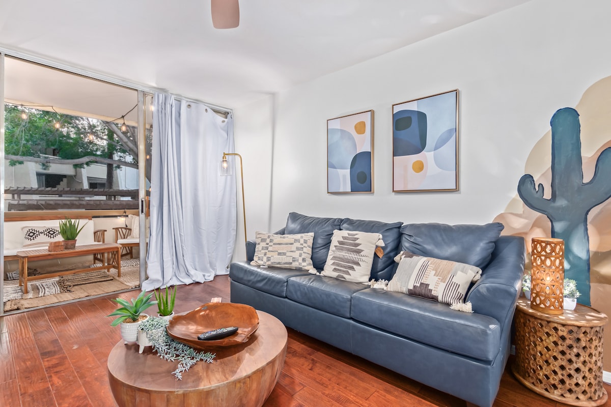 A cozy living area showcases a deep blue sofa adorned with decorative pillows. Two wall art pieces add color above the sofa, and natural light filters through a partially open sliding door leading to a patio. A wooden coffee table sits in front, surrounded by inviting decor.