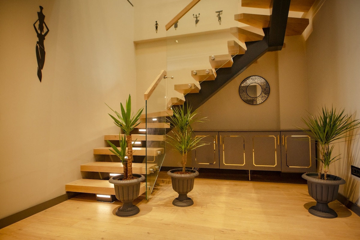 A well-lit entryway features wooden stairs leading to the upper floor. Decorative mirrors and wall art enhance the space. Potted plants are placed symmetrically along the walls, adding a touch of greenery to the modern design.