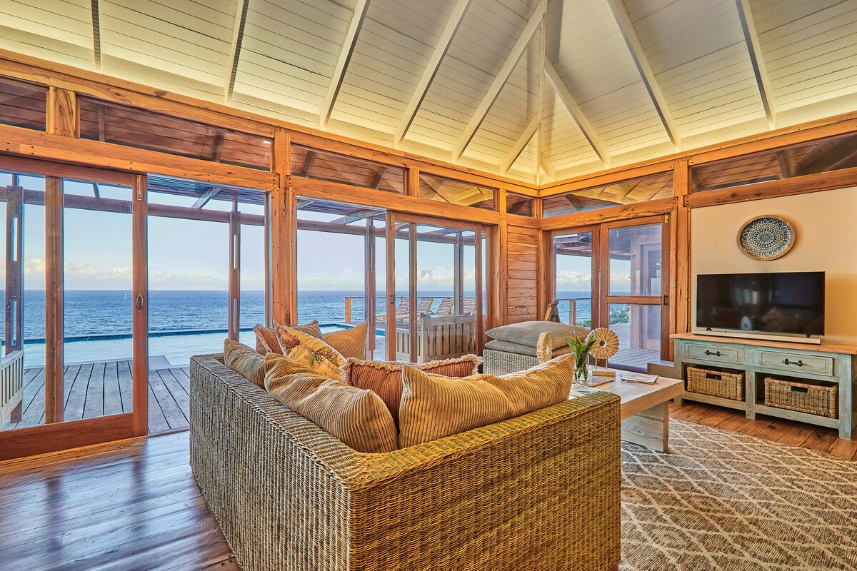 A spacious living area features a woven sofa adorned with cushions, surrounded by large windows that capture ocean views. Natural light fills the room, highlighting the wooden beams of the high ceiling. A flat-screen TV is positioned against the wall, complemented by a decorative piece above.