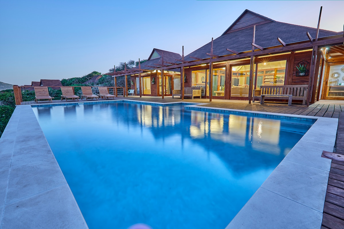 A serene outdoor area features a rectangular swimming pool, reflecting the clear sky. Sun loungers are lined up along the poolside, and wooden deck seating is positioned nearby. The villa's large windows invite views of the natural surroundings, enhancing the space's connection to nature.