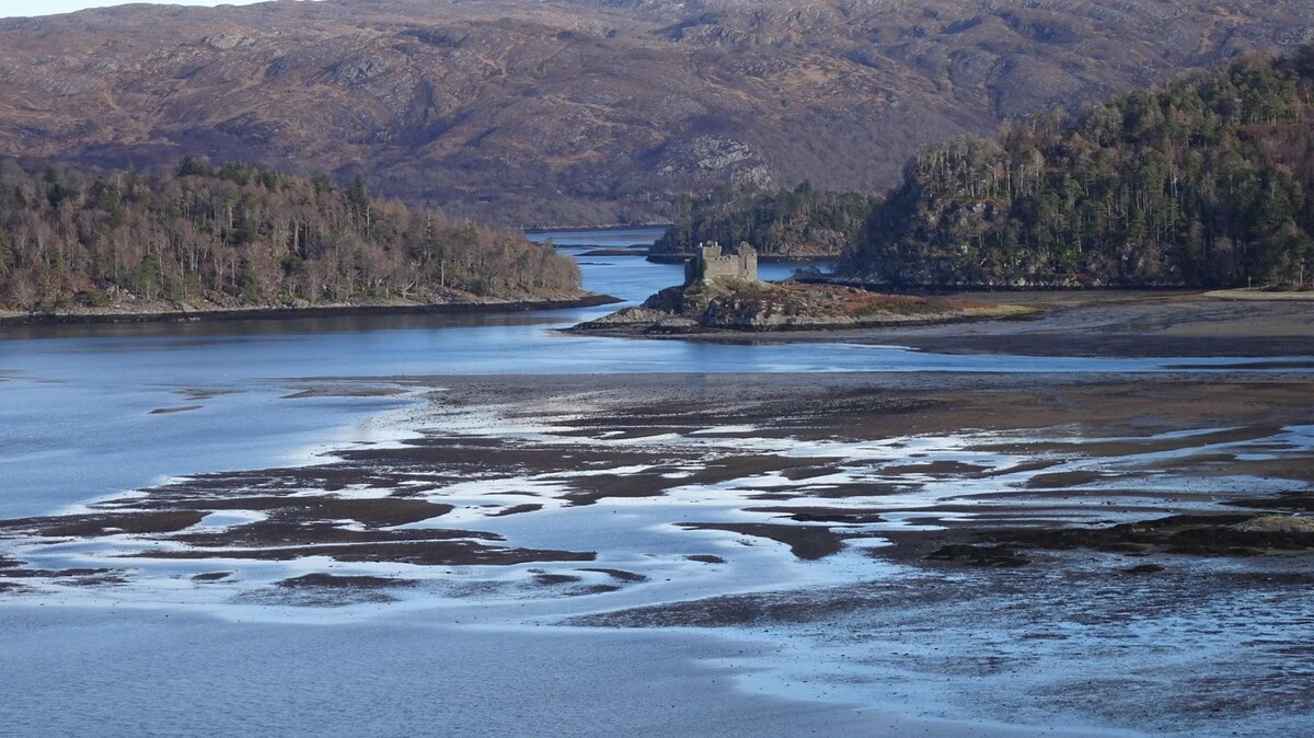 A serene view of the River Shiel showcases its winding waters surrounded by rugged hills, with a historic structure positioned on an island. The landscape features varying shades of brown and green, reflecting the natural beauty of the area.