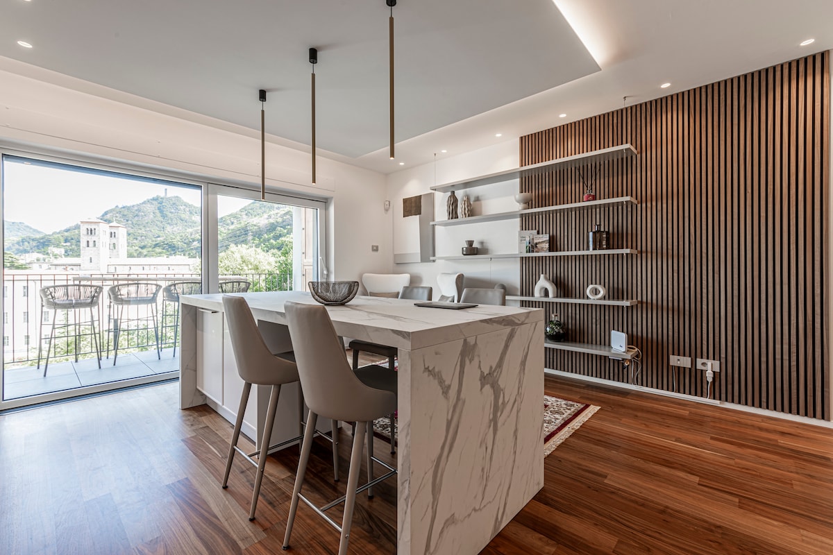 A modern dining area features a large marble table surrounded by sleek chairs. Open shelving displays decorative items, while natural light floods the space through a large glass door that opens to a balcony with views of the surrounding landscape.