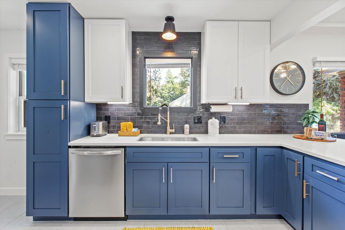 A modern kitchen features blue cabinetry paired with white upper cabinets. A stainless steel refrigerator is included, along with a sink and countertop workspace. Natural light enters through a window above the sink, and subtle decorative items enhance the space's functionality.